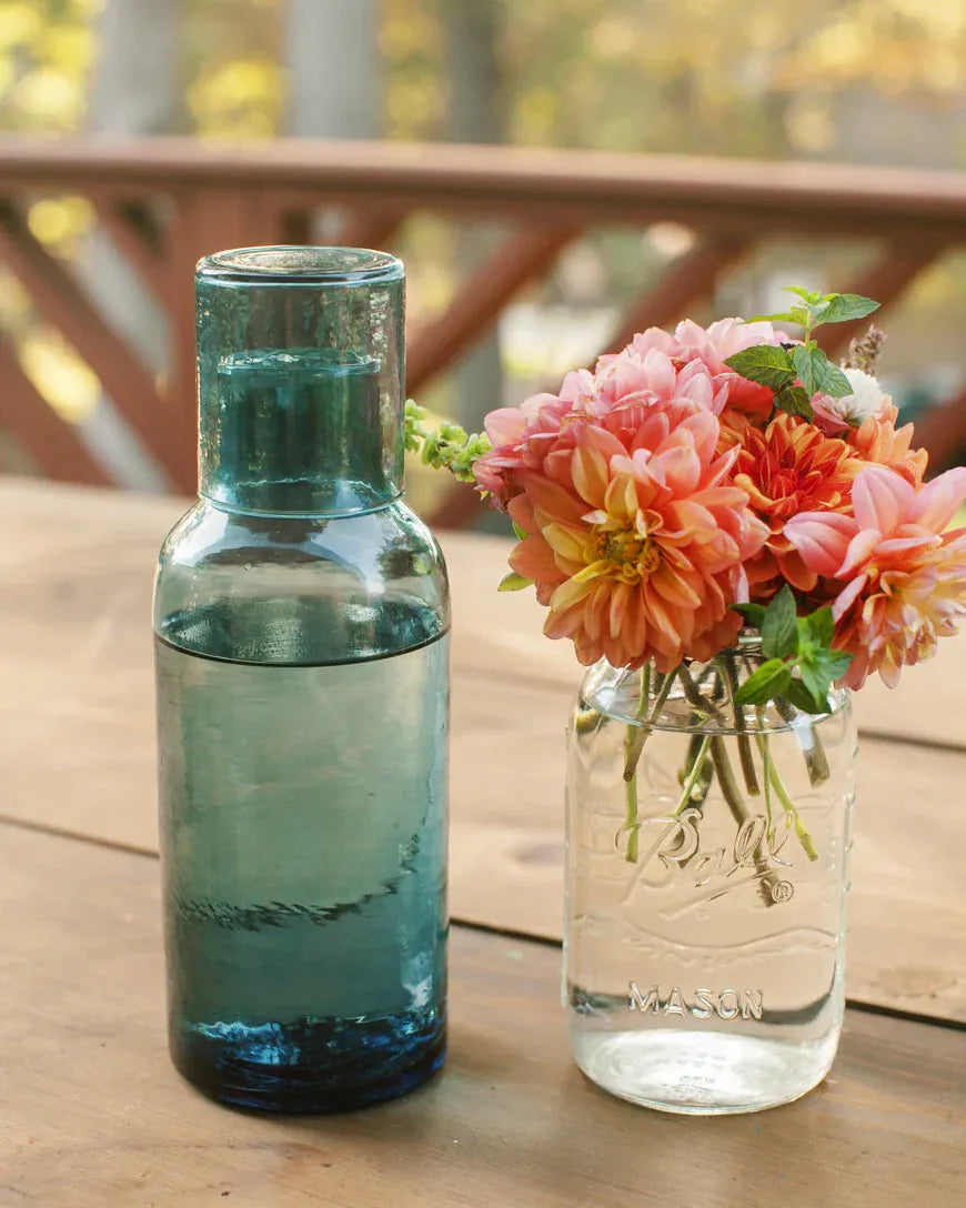 Transmutation-Carafe Glass Carafe - Hand blown 100% recycled blue glass set on an outdoor table next to a vase of flowers from the garden.